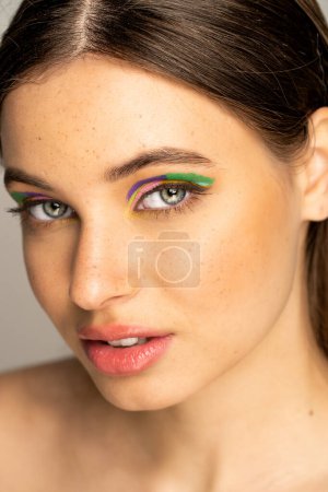 Teen model with colorful makeup and freckles looking at camera isolated on grey 