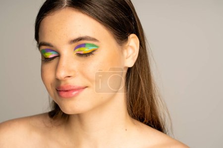 Pleased teenager with colorful eyeshadows and freckles posing isolated on grey 