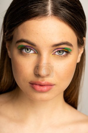 Téléchargez les photos : Portrait of freckled teenager with multicolored makeup standing isolated on grey - en image libre de droit