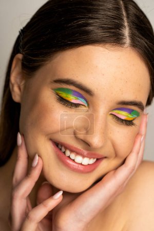 Pleased teen girl with colorful makeup touching face isolated on grey 