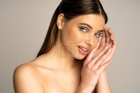 Smiling teen model with colorful makeup posing and looking at camera isolated on grey 