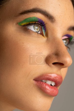 Close up view of teen model with freckles and colorful makeup looking away isolated on grey 