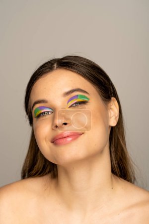 Téléchargez les photos : Joyful teen model with multicolored makeup looking at camera isolated on grey - en image libre de droit