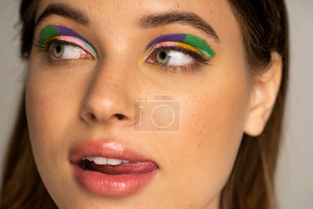 Close up view of teen model with multicolored visage sticking out tongue isolated on grey 