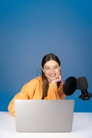 Téléchargez les photos : Cheerful teenager in orange jacket smiling at camera near laptop and studio microphone isolated on blue - en image libre de droit