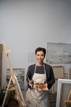 Photo for Young artist holding palette with paints and looking at camera near easels in workshop - Royalty Free Image