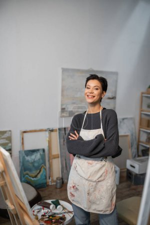 Téléchargez les photos : Cheerful artist in apron looking at camera near paints and easel in studio - en image libre de droit