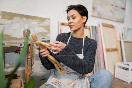 Artist in apron holding wooden doll near blurred paintings in studio 