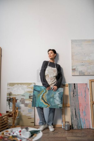Brunette artist in apron holding drawing near blurred paints in workshop 