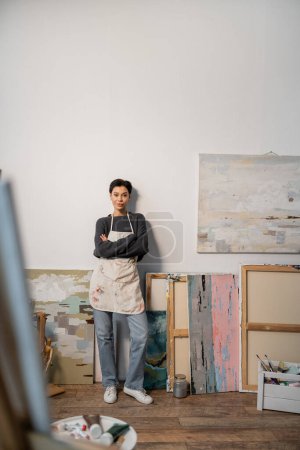 Young artist in apron crossing arms while standing near paintings in studio 