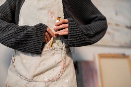 Téléchargez les photos : Cropped view of artist putting paintbrush in pocket on apron in studio - en image libre de droit