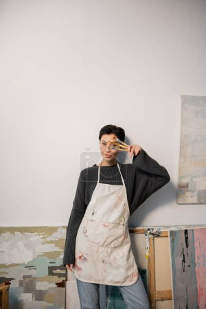 Brunette artist in apron holding paintbrushes near face and looking at camera near paintings in studio 