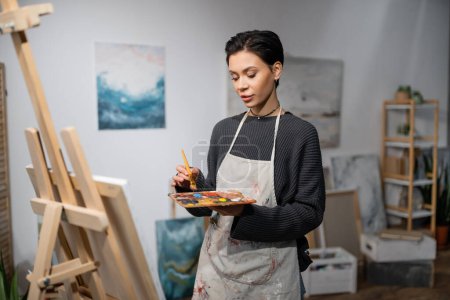 Young artist in apron holding paintbrush and palette near canvas on easel 