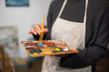 Cropped view of blurred artist in apron holding palette and paintbrush in studio 