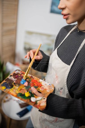 Cropped view of artist in apron mixing paints on palette in workshop 