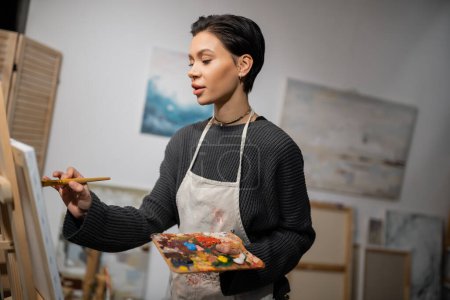 Brunette artist in apron painting on canvas in workshop 