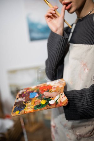 Cropped view of artist in apron holding palette and blurred paintbrush in studio 