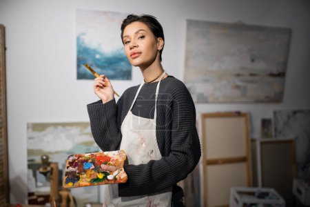 Téléchargez les photos : Brunette artist looking at camera while holding paintbrush and palette in blurred workshop - en image libre de droit