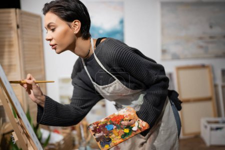 Téléchargez les photos : Young artist in apron painting on canvas in blurred studio - en image libre de droit