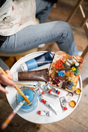 Téléchargez les photos : Top view of artist in apron putting paintbrush in jar with water near paints on table in workshop - en image libre de droit
