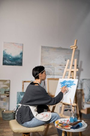 Téléchargez les photos : Side view of artist in apron painting on canvas in studio - en image libre de droit