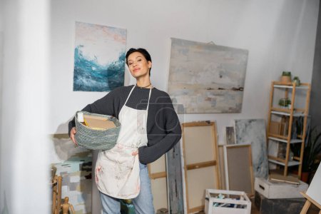 Short haired artist in apron holding basket with equipment near drawings in studio 