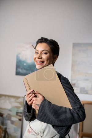 Portrait of cheerful artist in apron looking at camera while holding sketchbook 