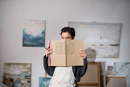 Brunette artist in dirty apron holding sketchbook near face in studio 