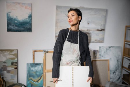 Artist in dirty apron holding sketchbook and looking away near blurred drawings in workshop 