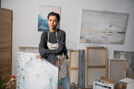 Téléchargez les photos : Young artist looking at camera while standing near painting in studio - en image libre de droit