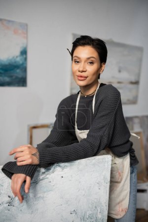 Téléchargez les photos : Brunette artist in dirty apron looking at camera while standing near painting in studio - en image libre de droit
