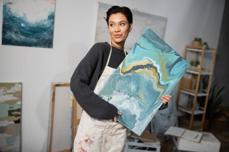 Foto de Pleased artist in apron looking away while holding painting in workshop - Imagen libre de derechos