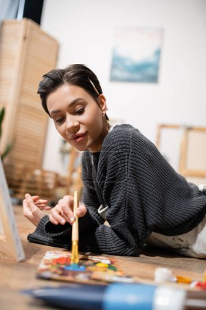 Téléchargez les photos : Brunette artist in sweater mixing paints on palette while lying on floor - en image libre de droit