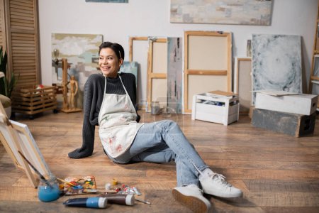 Positive artist in apron sitting on floor near paints and painting in workshop 