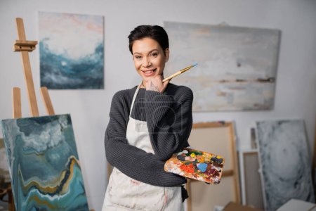 Cheerful tattooed artist holding paintbrush and palette in studio 