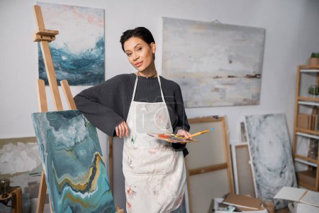 Foto de Brunette artist holding paintbrush and palette near canvas on easel in studio - Imagen libre de derechos