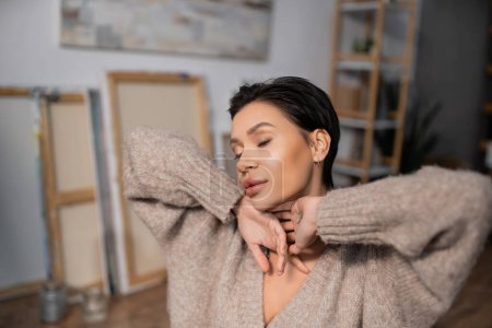 Brunette artist in sweater closing eyes in blurred workshop 