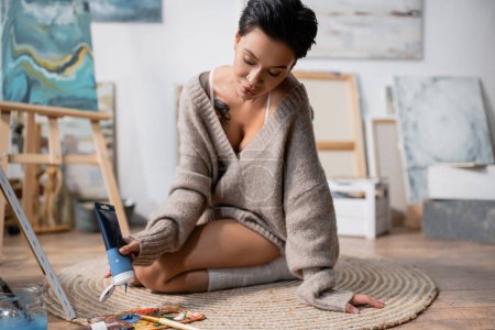 Artist in sweater pouring paint on palette on floor in workshop 