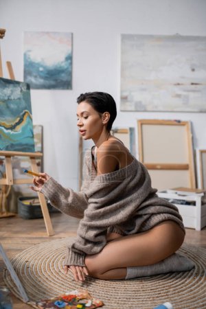 Sexy artist holding paintbrush near palette on floor in studio 