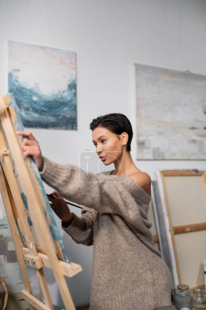 Sexy artist in sweater painting on canvas in workshop 