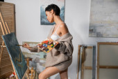 Side view of seductive artist in sweater painting on canvas and easel in studio  Poster #634326078