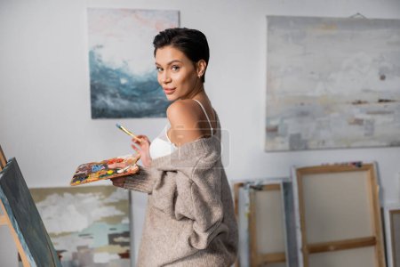 Foto de Brunette artist in bra and sweater holding palette with paints near canvas in workshop - Imagen libre de derechos