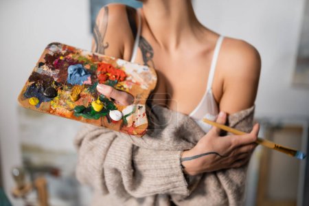 Cropped view of blurred tattooed artist in bra and sweater holding paintbrush and palette in workshop 