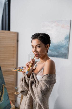 Sensual artist holding paintbrush near lips and looking at camera in studio 