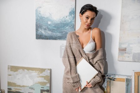 Sexy artist with pencil and sketchbook standing near paintings in studio