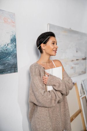 Smiling and sexy artist in sweater with pencil and sketchbook standing in workshop 