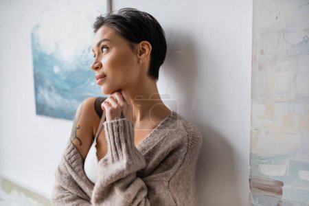 Seductive artist in sweater and lingerie standing near paintings on wall 