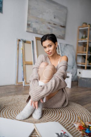 Sensual brunette artist in sweater and socks sitting near sketchbook and palette in workshop 