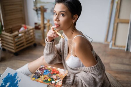 Sexy artist in sweater and bra holding paintbrush and palette in workshop 