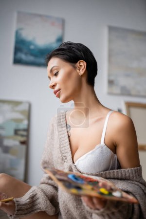 Young sexy artist in bra and sweater holding blurred palette in workshop 